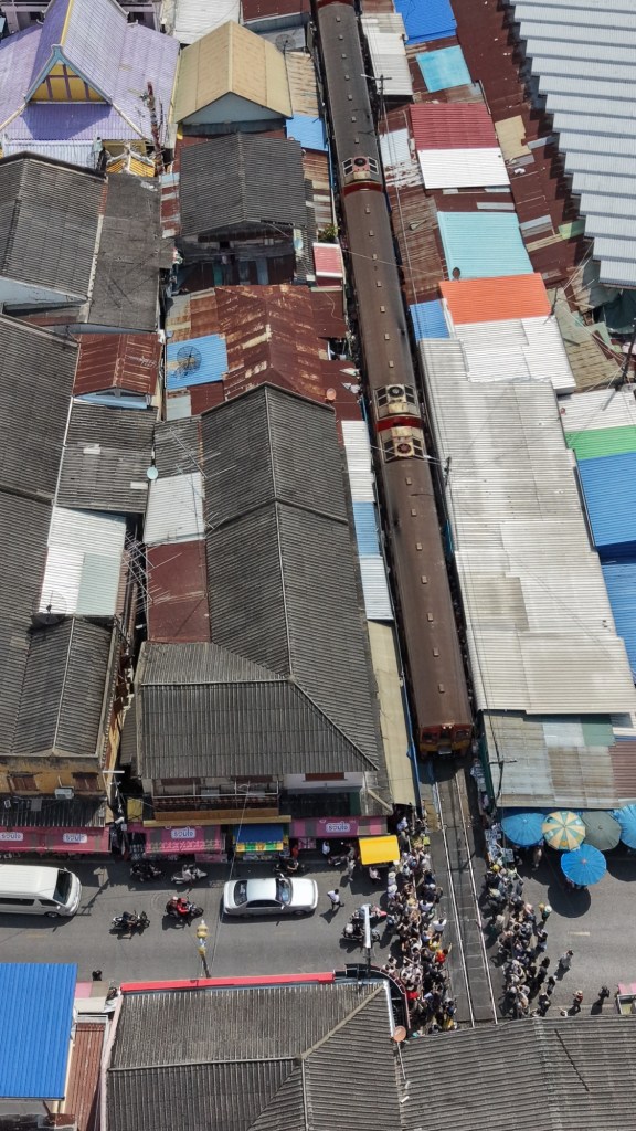 Aerial view of Mae Klong Railway Market in Thailand with a train running through the center of the market and crowds gathered along the tracks
