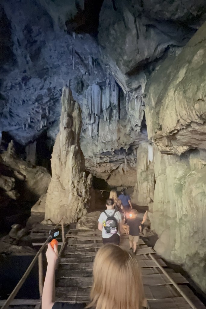 Visitors walking along wooden pathways inside Tham Lod Cave near Pai, Thailand while a guide lights the cave with a lantern.