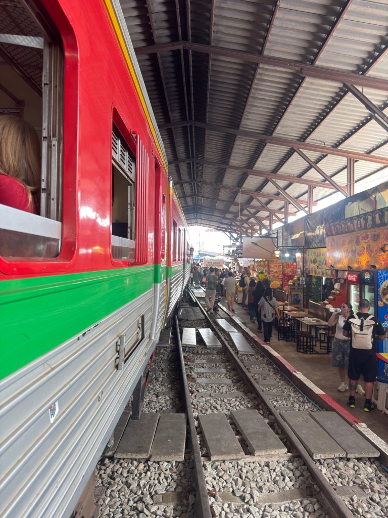 Train passing through Mae Klong Railway Market in Thailand with vendors and visitors standing along the tracks
