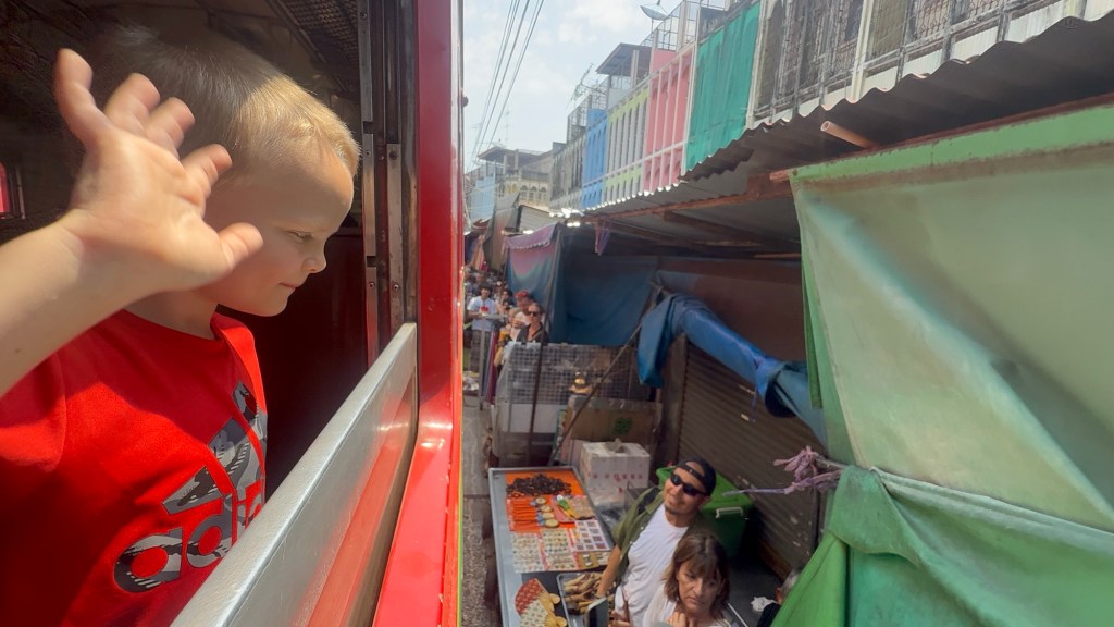 Child leaning out of train window as it passes through Mae Klong Railway Market with vendors and visitors standing close to the tracks