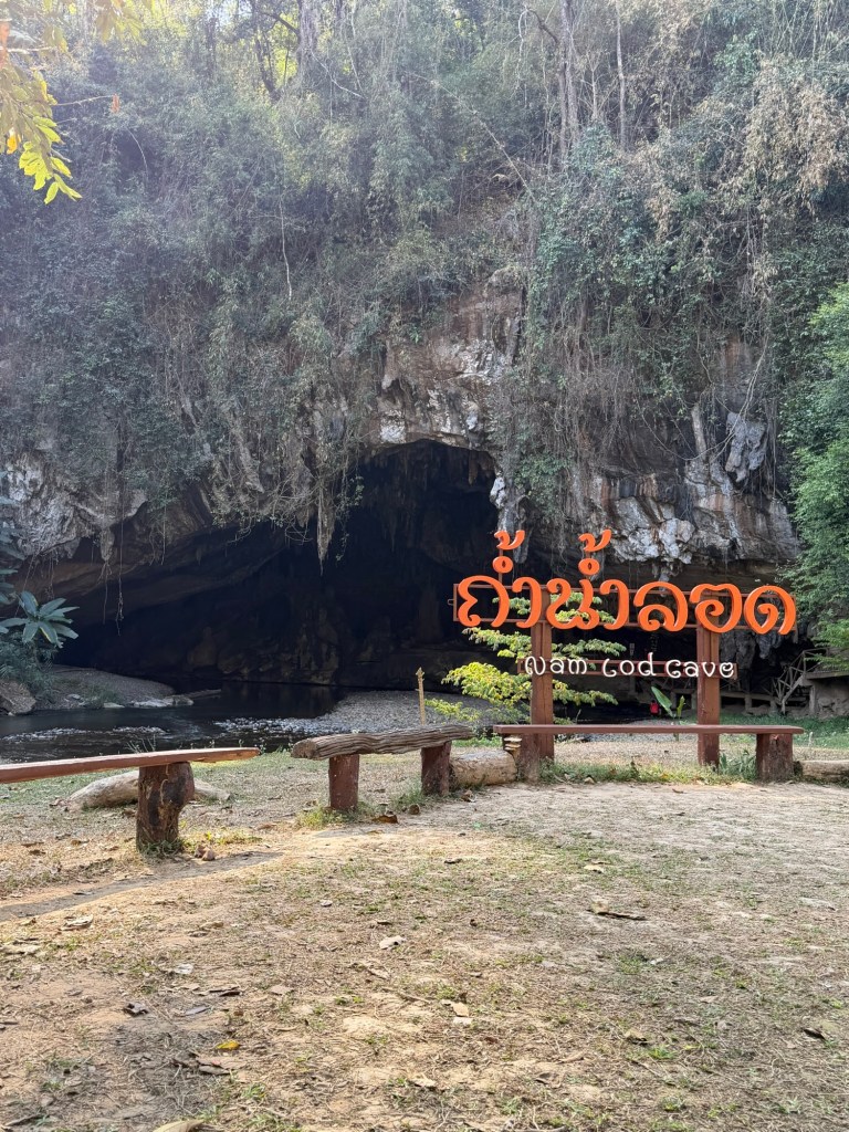 Entrance to Tham Lod Cave near Pai, Thailand, with the Lod Cave sign and river flowing into the large limestone cave surrounded by jungle.